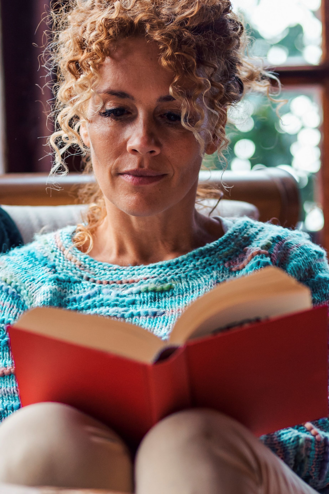 A woman reading an open book while curled up in a chair by a bright window